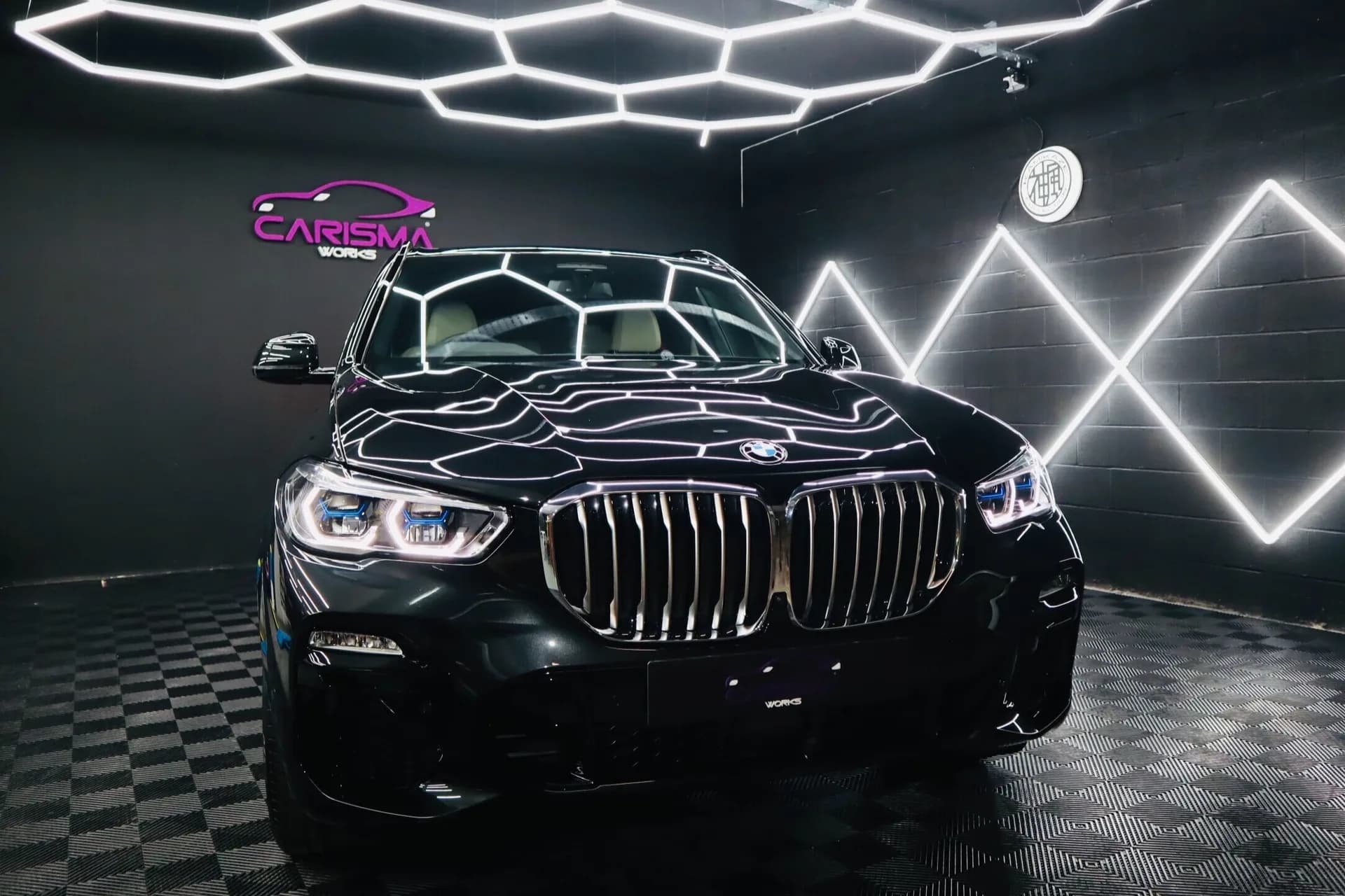 Black BMW X5 shown front three-quarter inside the Carisma Works studio, branded neon signage visible behind it
